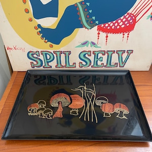 May include: A rectangular black tray with a glossy finish, featuring a decorative design of stylized brown and gold mushrooms and tall grass. The tray is set against a backdrop of a vintage poster with the text "SPIL SELV".