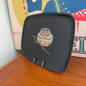 May include: Black square tray with rounded corners. The tray features a decorative design of a vase with deer and geometric patterns, along with a bundle of wheat. The tray has two silver metal hooks.