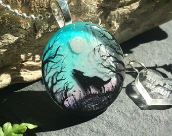 May include: A round pendant necklace with a silver chain. The pendant features a wolf howling at the moon in a forest scene, with a teal and gray background. A small heart-shaped charm hangs next to the pendant.