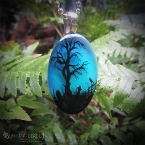 May include: A blue glass pendant with a black silhouette of a tree, a bird, and a fence. The pendant is on a silver chain.