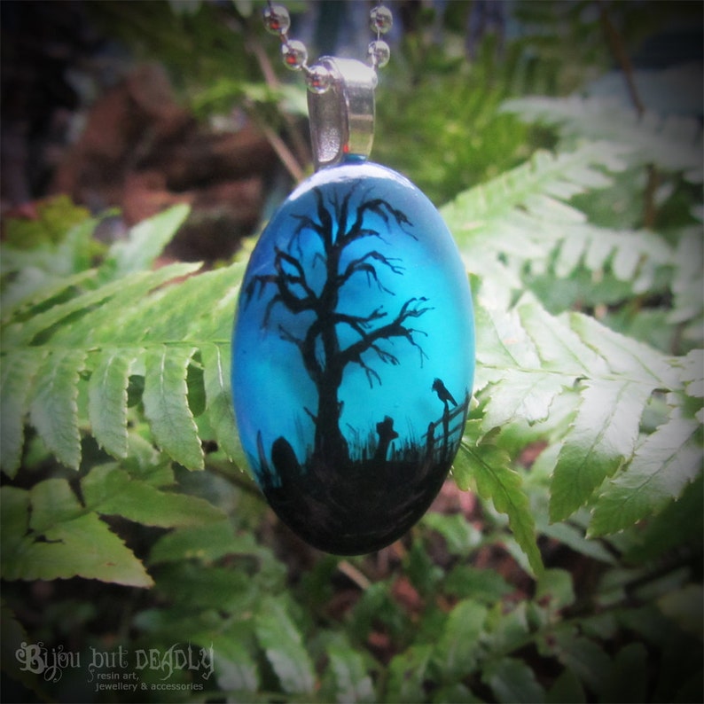 May include: A blue glass pendant with a black silhouette of a tree, a bird, and a fence. The pendant is on a silver chain.