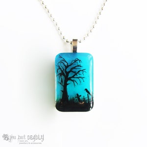 May include: A blue glass pendant necklace with a black silhouette of a tree, two birds, and a tombstone. The pendant is on a silver chain.