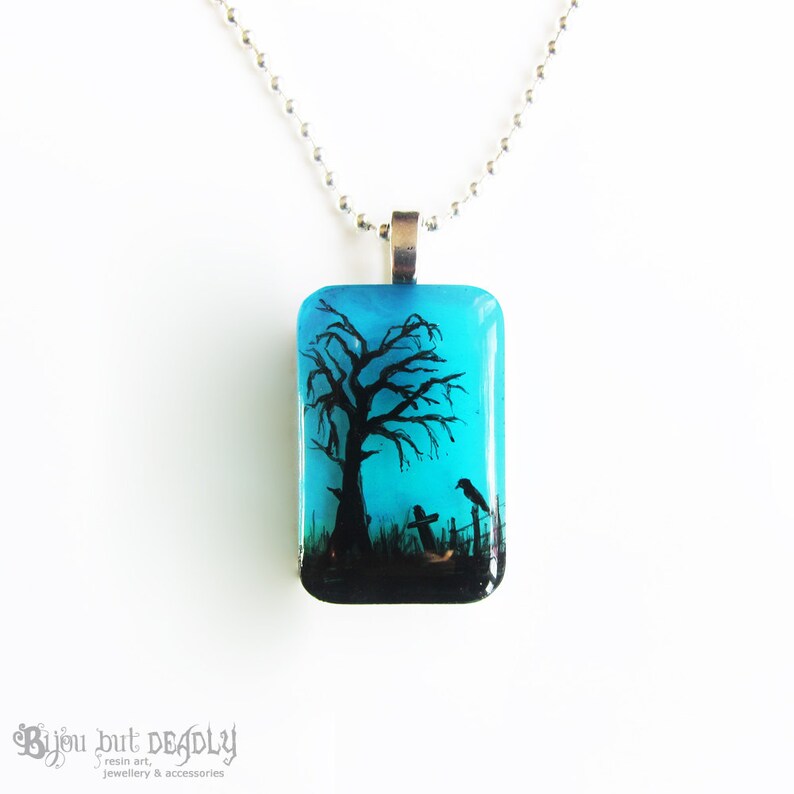 May include: A blue glass pendant necklace with a black silhouette of a tree, two birds, and a tombstone. The pendant is on a silver chain.