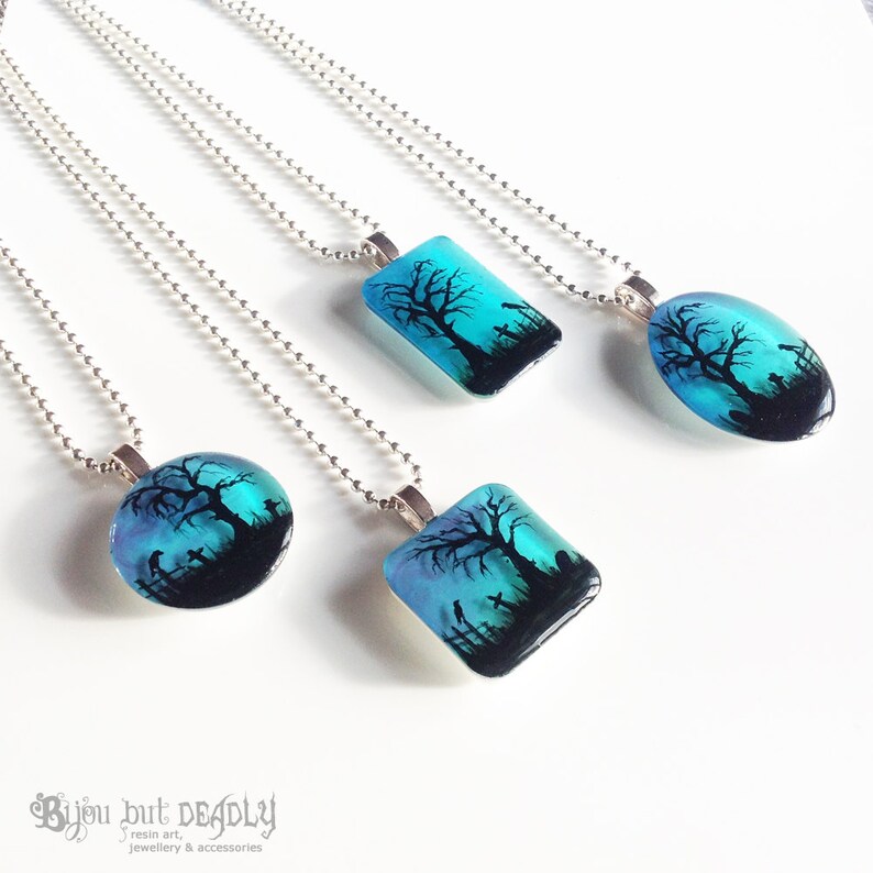 May include: Four silver necklaces with blue and black resin pendants featuring a silhouette of a tree and a graveyard. The pendants are in different shapes, including round, oval, and square.