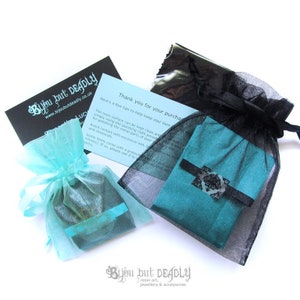 May include: An overhead shot of jewelry packaging. Two small teal fabric pouches are tied with teal ribbons and placed in sheer turquoise and black bags. A black card with white text and a blue card with text are also visible. The brand name "Bijou but Deadly" is printed on the cards.