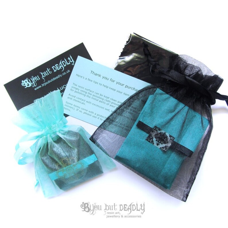 May include: An overhead shot of jewelry packaging. Two small teal fabric pouches are tied with teal ribbons and placed in sheer turquoise and black bags. A black card with white text and a blue card with text are also visible. The brand name "Bijou but Deadly" is printed on the cards.