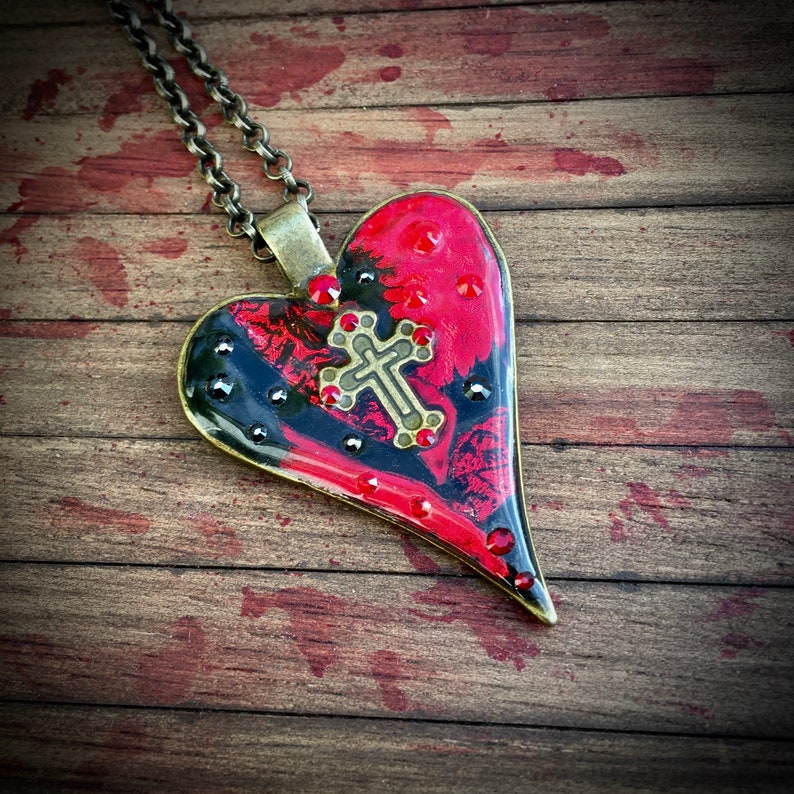 May include: A heart-shaped pendant necklace with a bronze chain. The pendant features a red and black design with a cross and red and black embellishments. The necklace is on a wooden surface.