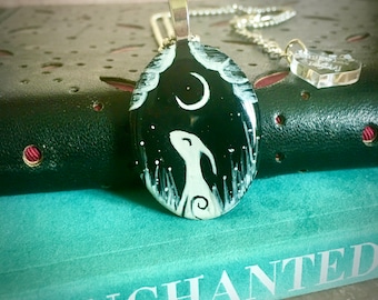 May include: A silver chain necklace with a black and white painted pendant featuring a white rabbit and a crescent moon. The pendant is oval shaped and has a white background with black details. The necklace is on a book with the word "ENCHANTED" printed on the cover.