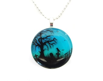 May include: A round pendant necklace with a spooky graveyard scene. The pendant features a black tree, a crow, and tombstones against a teal and blue gradient background. The necklace has a silver chain.