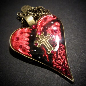 May include: A heart-shaped pendant necklace with a bronze chain. The pendant features a red and black design with a gold cross and red rhinestones. The pendant has a bronze border.