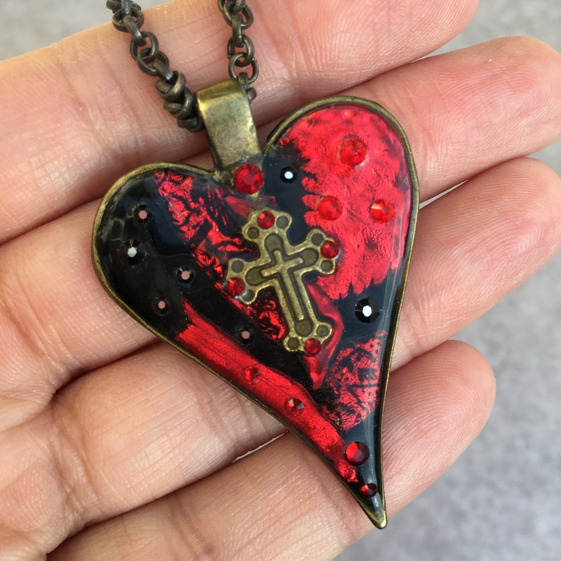 May include: A heart-shaped pendant necklace with a red and black design. The pendant features a brass cross and is adorned with red and black crystals. The necklace has a dark chain.