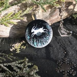 May include: A round pendant with a silver bail features a white hare leaping across a dark night sky with a full moon. The pendant has a black background with white and teal accents. A small heart-shaped charm with text hangs from a chain.