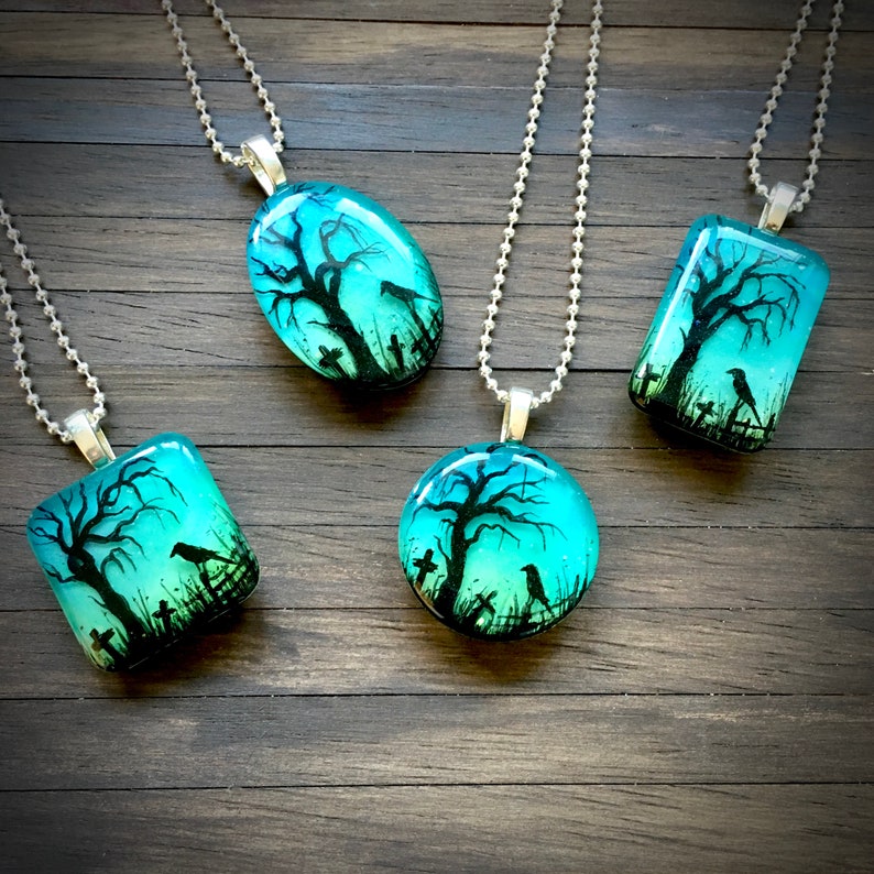 May include: Four glass pendants on silver chains, each featuring a dark silhouette of a tree with crows and a graveyard against a teal and blue gradient background.