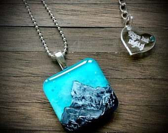 May include: A silver chain necklace with a square glass pendant featuring a painted mountain scene in shades of blue and grey. The pendant is attached to a silver chain with a heart-shaped charm that reads "You just desigly".