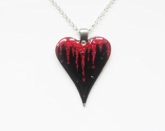 May include: A silver chain necklace with a black and red heart pendant. The heart is dripping with red glitter and has a small silver rhinestone in the center.