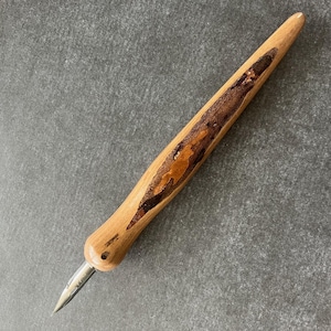 May include: A wooden calligraphy pen with a light brown handle and a dark brown, textured inlay. The pen features a silver nib and is designed for writing or drawing. The pen is angled on a gray surface.
