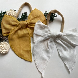 May include: Two fabric hair bows, one in a mustard yellow color and one in white. Both bows are tied in a large, floppy bow shape with long tails.