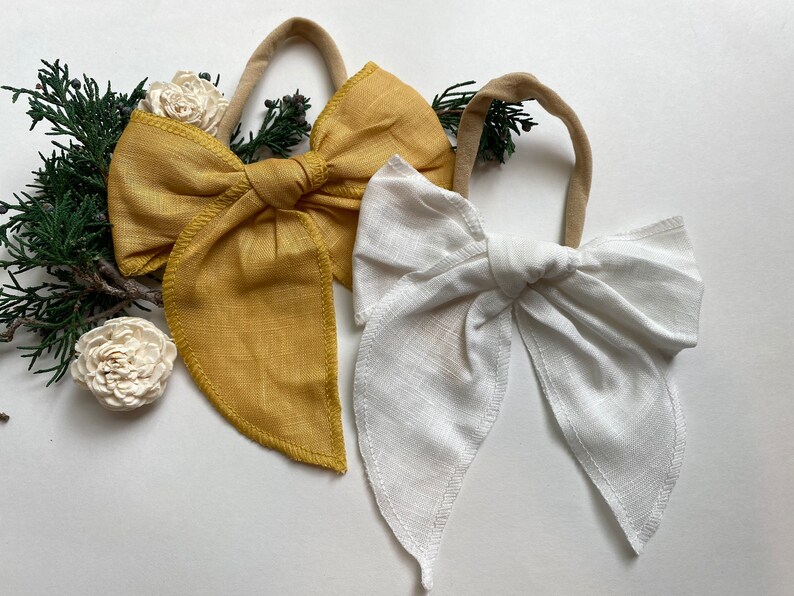 May include: Two fabric hair bows, one in a mustard yellow color and one in white. Both bows are tied in a large, floppy bow shape with long tails.