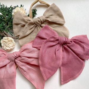 May include: Three fabric hair bows in different colors. The bows are made of a soft, linen-like material. The colors are beige, pink, and a darker shade of pink.