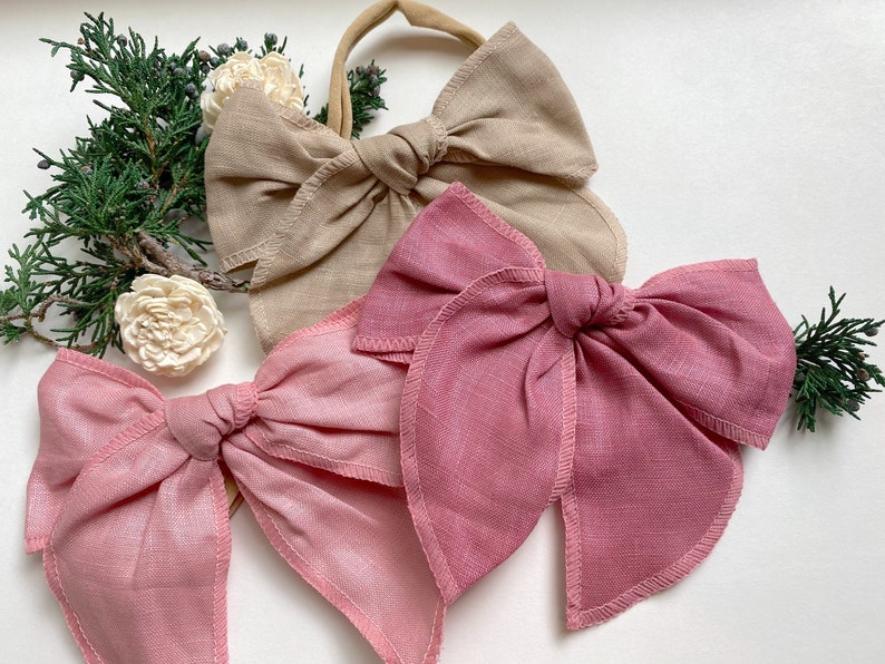 May include: Three fabric hair bows in different colors. The bows are made of a soft, linen-like material. The colors are beige, pink, and a darker shade of pink.