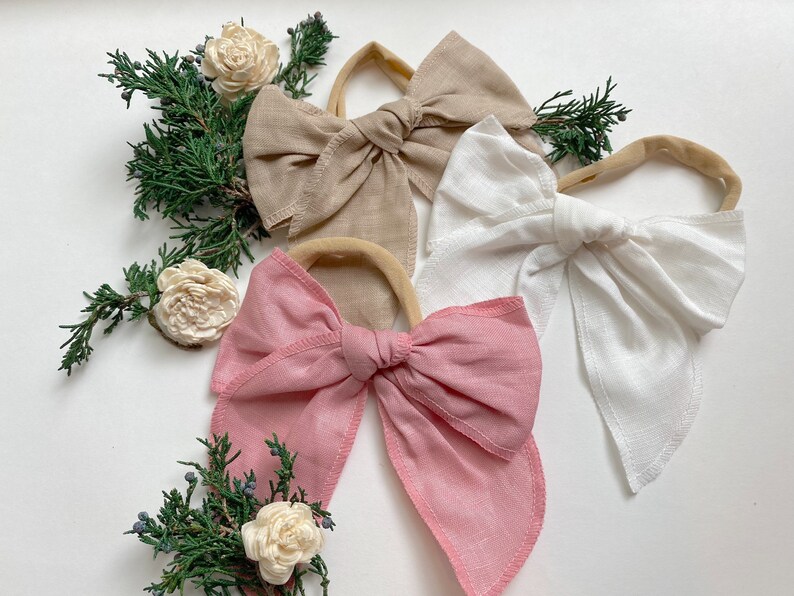 May include: Three fabric hair bows with elastic bands. The bows are in shades of pink, beige, and white.