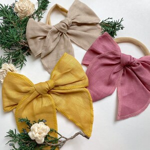 May include: Three fabric hair bows in different colors: beige, mustard yellow, and dusty rose. Each bow is tied with a knot in the center and has a simple, elegant design.
