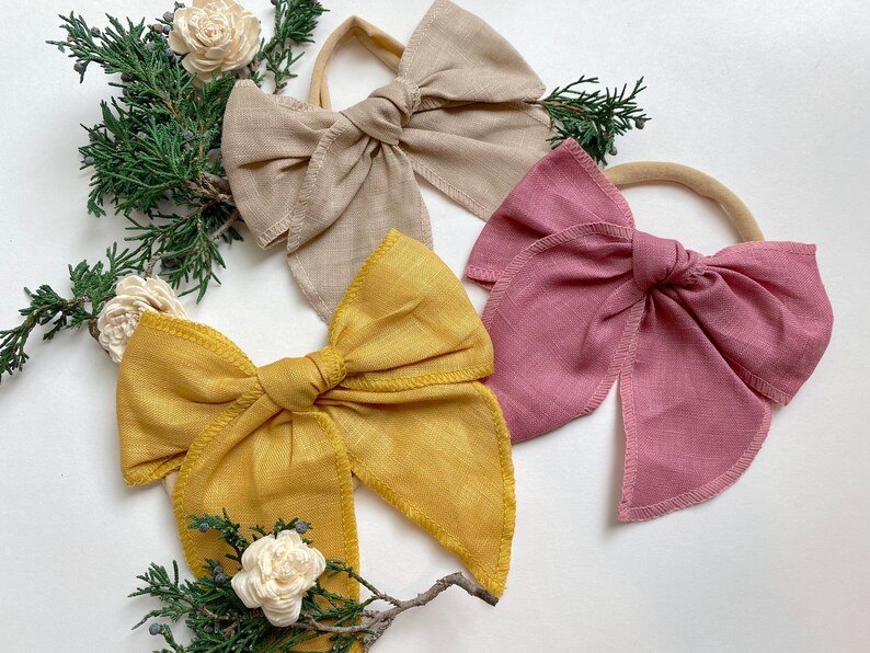 May include: Three fabric hair bows in different colors: beige, mustard yellow, and dusty rose. Each bow is tied with a knot in the center and has a simple, elegant design.