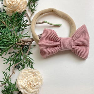 Puede incluir: Un lazo de pana rosa atado a una diadema el&aacute;stica de color nude. La diadema est&aacute; sobre un fondo blanco con una planta verde.