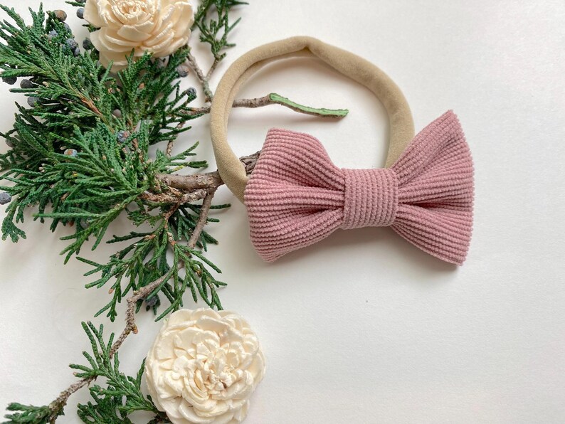 Puede incluir: Un lazo de pana rosa atado a una diadema el&aacute;stica de color nude. La diadema est&aacute; sobre un fondo blanco con una planta verde.