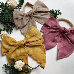 May include: Three fabric hair bows in different colors: beige, mustard yellow, and dusty rose. The bows are tied with a simple knot and have a frayed edge. The bows are on a white background.
