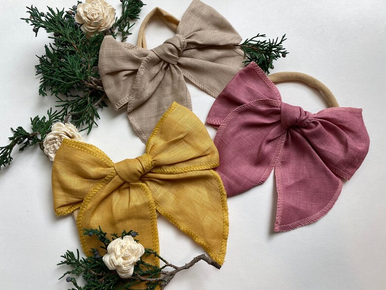 May include: Three fabric hair bows in different colors: beige, mustard yellow, and dusty rose. The bows are tied with a simple knot and have a frayed edge. The bows are on a white background.