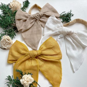 May include: Three fabric headbands with large bows. The bows are in beige, white, and mustard yellow.