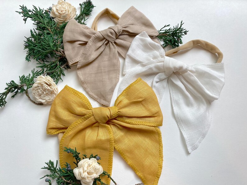 May include: Three fabric headbands with large bows. The bows are in beige, white, and mustard yellow.