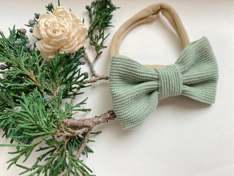 Puede incluir: Un lazo de pana verde salvia atado a una diadema el&aacute;stica nude. La diadema est&aacute; sobre una superficie blanca con una rama de vegetaci&oacute;n.