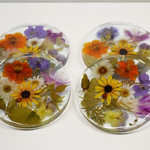 May include: Set of four clear resin coasters, each embedded with a vibrant assortment of dried flowers. The coasters feature orange, yellow, purple, and white blooms, along with green leaves, creating a decorative and functional item.