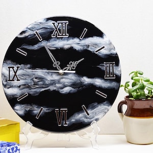 May include: A round wall clock featuring a black background with swirling white marble-like patterns. The clock face displays silver Roman numerals at the twelve, three, six, and nine o'clock positions, with silver hour, minute, and second hands.