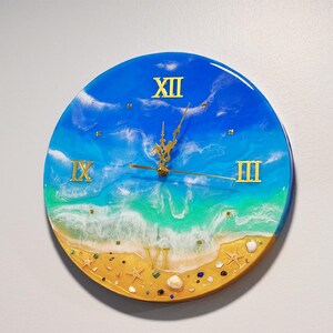 May include: Round wall clock with a beach-themed design. The clock face features a blue and white ocean scene with gold Roman numerals and clock hands. The bottom of the clock has a sandy beach design with seashells and starfish.