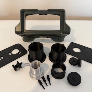 May include: An assortment of black and silver mechanical parts, including a rectangular frame, cylindrical components, and various small pieces. The frame is olive green. The parts are arranged on a white surface.