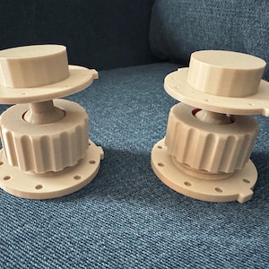 May include: Two beige, 3D-printed objects with a unique design. Each has a circular base with holes, a ridged midsection, a flat disc, and a cylindrical top. The objects are displayed on a blue fabric surface.