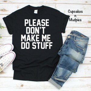 May include: Black t-shirt with the phrase "PLEASE DON'T MAKE ME DO STUFF" in bold white letters. The shirt is paired with blue jeans and white sneakers. The image is on a light wood background.