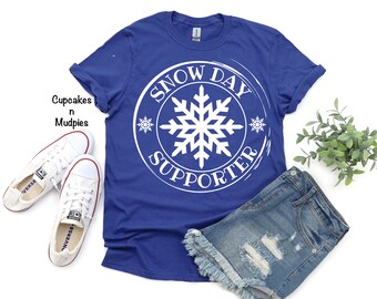 May include: A royal blue short-sleeve t-shirt featuring a large white circular graphic with a central snowflake and the text "SNOW DAY SUPPORTER". The shirt is displayed alongside a pair of white canvas sneakers and distressed denim shorts.