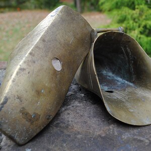 May include: Two antique brass horse shoeing tools. The tools are made of brass and have a worn, aged patina. The tools are shaped like a scoop and a flat piece of metal with a hole in the center.