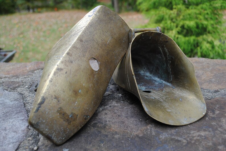May include: Two antique brass horse shoeing tools. The tools are made of brass and have a worn, aged patina. The tools are shaped like a scoop and a flat piece of metal with a hole in the center.