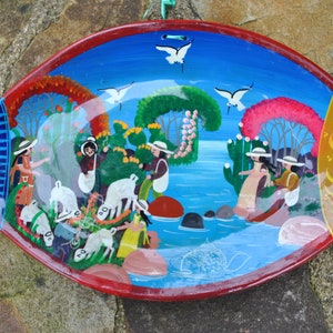 Peut inclure: Une assiette décorative peinte à la main en forme de poisson, avec une bordure rouge. L'intérieur présente une scène colorée avec des personnages, des moutons, des arbres et des oiseaux sur fond de ciel bleu et d'eau. La queue du poisson est bleue et la tête est jaune.