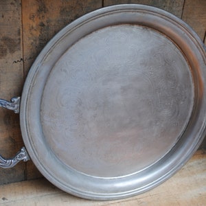 Puede incluir: Una bandeja de servir plateada con diseños ornamentados y dos asas. La bandeja está hecha de metal y tiene un acabado brillante.