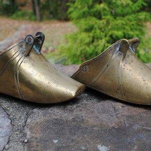 May include: Two antique brass stirrups with ornate designs. The stirrups are made of a smooth, polished brass and have a slightly curved shape.