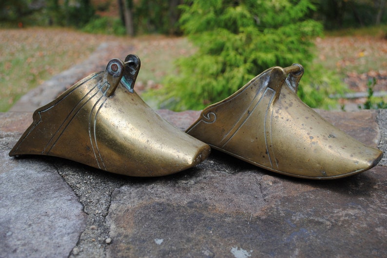 May include: Two antique brass stirrups with ornate designs. The stirrups are made of a smooth, polished brass and have a slightly curved shape.