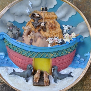 May include: A decorative, round Noah's Ark figurine. The ark is red and blue, filled with pairs of animals, including camels, zebras, and cows. Two walruses and two rabbits stand at the base. The background features a stormy sky.