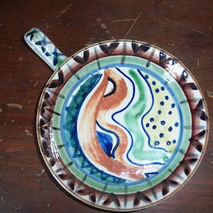 May include: A small, white ceramic dish with a handle. The dish is decorated with a colourful, abstract design in shades of blue, green, yellow, and orange.