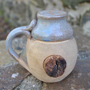 May include: A ceramic mug with a light blue glaze and a wooden stopper. The mug has a handle and a round body. The wooden stopper is in the shape of a circle and is attached to the side of the mug.